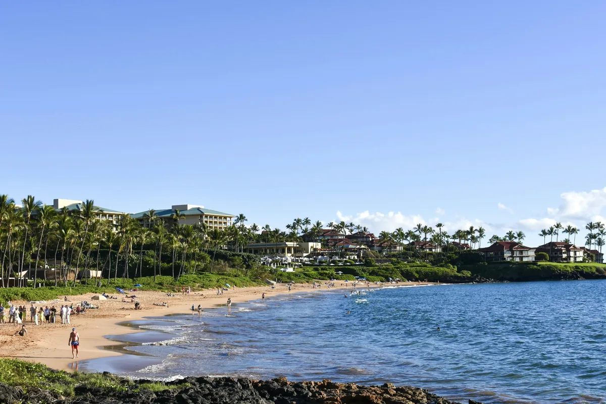 A curve of Maui beach with palms and low resort buildings along the shore under a blue sky